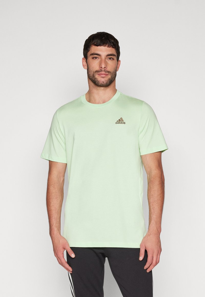 Camiseta de algodón verde claro con cuello redondo, que presenta un logo de adidas en marrón en el lado izquierdo del pecho. Mangas cortas y un ajuste relajado.