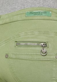 Green denim pants featuring a small zippered pocket, metal hardware, and branded tag. Smooth texture with contrasting stitching details.