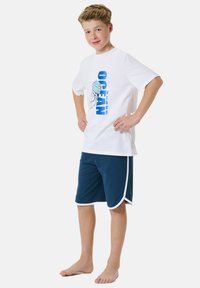 White cotton t-shirt with blue "OCEAN" graphic and jellyfish design. Navy shorts with white side accents. Model standing barefoot, smiling.