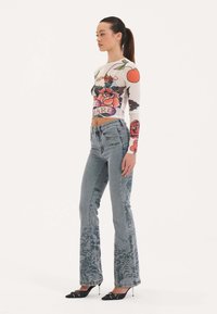 Langsleeve, doorzichtig top met bloemenprints in rood en groen; high-waisted, lichtblauwe jeans met ingewikkelde grijze bloemenontwerpen; zwarte hakken.
