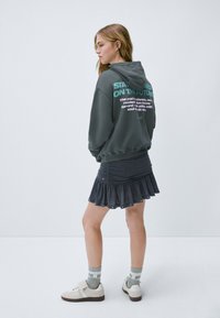 Sudadera gris con capucha y texto decorativo en la parte posterior, combinada con una falda oscura con dobladillo con volantes. La modelo lleva zapatillas ligeras y calcetines estampados.