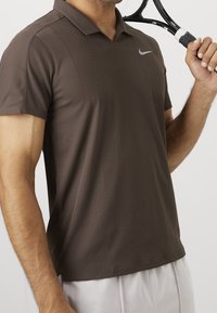 Homme portant un polo marron Nike tenant une raquette de tennis noire et blanche sur son épaule.