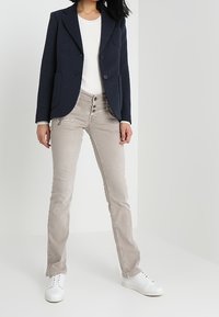 Blazer navy de dos botones y bolsillos, combinado con jeans slim-fit en beige claro con cremalleras y zapatillas blancas. Diseño simple y estructurado.