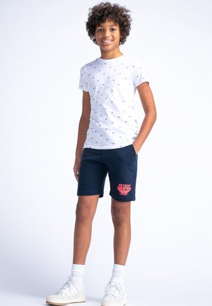 T-shirt blanc à manches courtes avec de petits motifs bleus, associé à un short bleu marine arborant un logo rouge. Baskets blanches et chaussettes courtes côtelées.