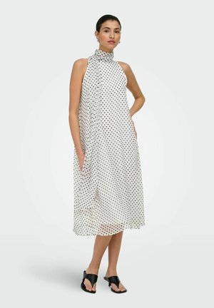 Femme portant une robe midi blanche sans manches à pois noirs, un lien haut au cou, et des sandales noires à talons ouverts, debout avec une main sur la hanche.