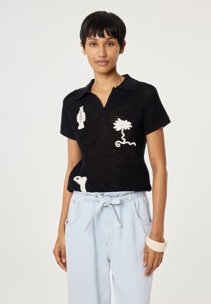 Frau trägt ein schwarzes, kurzes Netzoberteil mit weißen Blumenmustern, hellblaue, hoch Taillen gebundene Hose und ein weißes Armband.