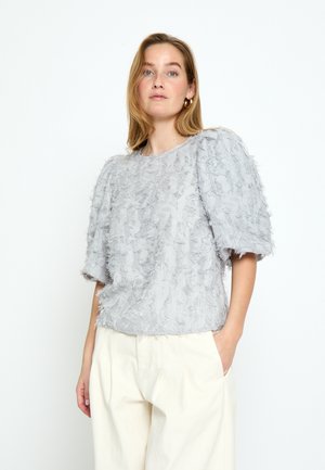 Vrouw draagt een lichtgrijze blouse met korte mouwen en een gestructureerde stof, en crème wijde broek, staand met één hand in de zak tegen een witte achtergrond.