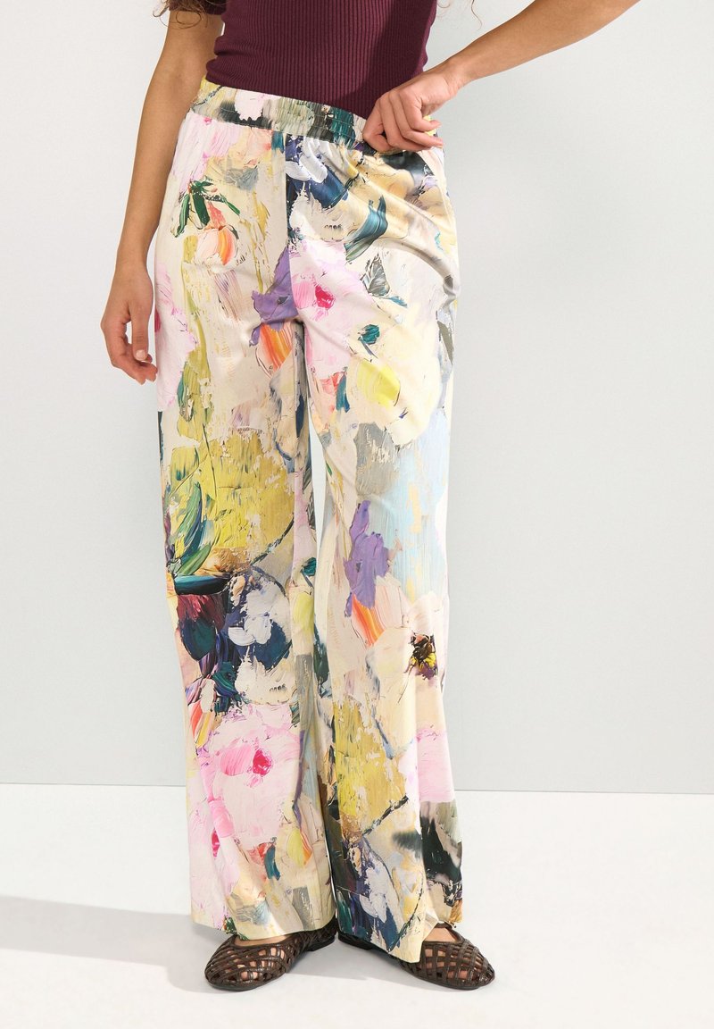 Femme portant un pantalon à jambes larges avec un imprimé floral abstrait coloré et des chaussures slip-on tissées foncées, debout devant un fond uni.