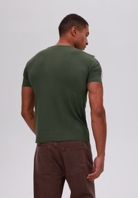 Camisa de manga corta verde oscuro con una textura suave. Presenta acentos blancos en el hombro, combinada con pantalones marrones con dos bolsillos traseros.
