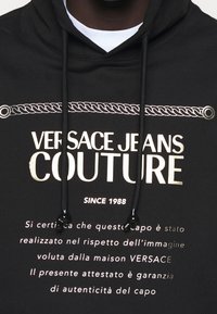 Black hoodie featuring "Versace Jeans Couture" logo in gold, with a decorative rope design above and below the text. Drawstrings with black accents.