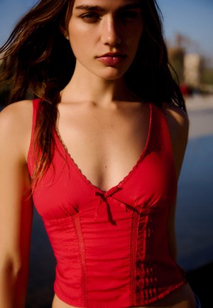 Mlada ženska z dolgimi lasmi, oblečena v rdečo brezrokavnik, stoji na prostem blizu vode z zamegljenimi stavbami v ozadju.