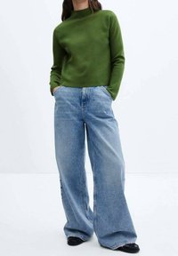 Groene trui met lange mouwen en hoge kraag, gecombineerd met lichtblauwe denim jeans met wijde pijpen. Gladde textuur met een ontspannen pasvorm. Zwarte schoenen.