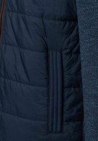 Marineblaue Steppjacke mit gesteppten Paneelen, einem strukturierten Strickärmel und einem kontrastierenden braunen Reißverschluss mit seitlicher Taschenverzierung.