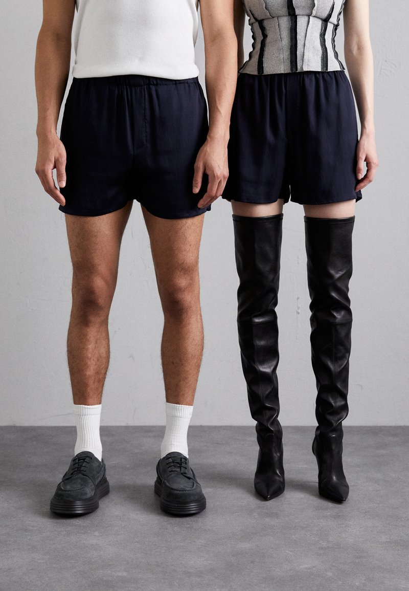 Eine Person trägt marineblaue Shorts, kombiniert mit einem weißen Tanktop und dunklen Schuhen; daneben steht eine zweite Person in hohen schwarzen Lederstiefeln und einem taillierten Oberteil.