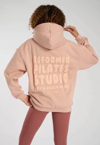 Rosa hoodie med en stor, upphöjd trycktext som säger "REFORMER PILATES STUDIO SOUTH BEACH MIAMI EST. 1953" på baksidan. Mjuk tyg, avslappnad passform.