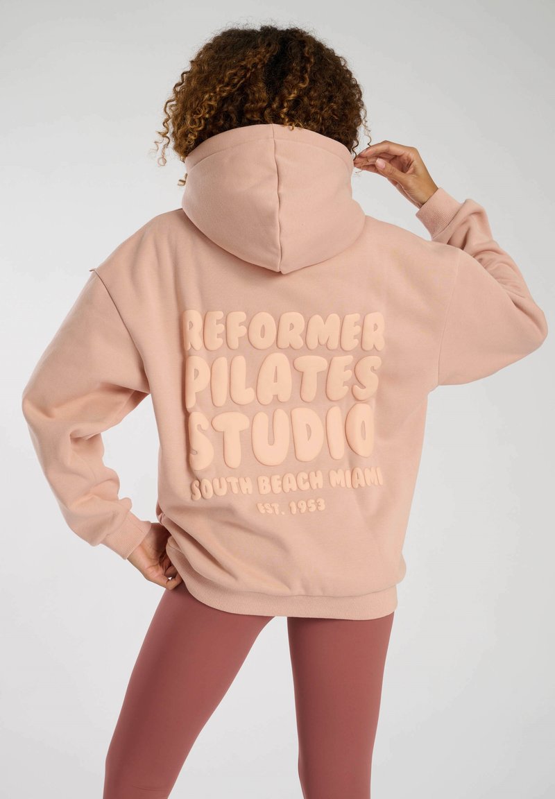Rosa hoodie med en stor, upphöjd trycktext som säger "REFORMER PILATES STUDIO SOUTH BEACH MIAMI EST. 1953" på baksidan. Mjuk tyg, avslappnad passform.