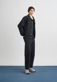 Jaqueta de ganga preta com botões, combinada com uma camisa preta e calças pretas. O calçado são ténis cinzentos. Sem texturas ou padrões notáveis.