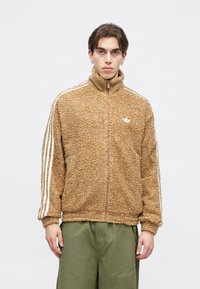 Καφέ, φουντωτή ζακέτα με φερμουάρ και ψηλό γιακά. Διαθέτει λευκές ρίγες κατά μήκος των μανικιών και ένα μικρό λογότυπο στο στήθος. Φοριέται με πράσινα παντελόνια.