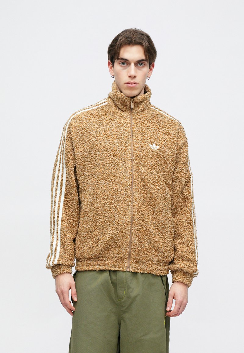 Brown, fluffy zip-up jacket with high collar. Features white stripes along sleeves and a small logo on the chest. Worn with green pants.