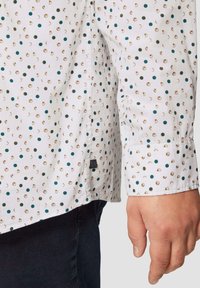 Witte shirt met een patroon van teal en gouden stippen, met een button-down kraag, lange mouwen en een gestikte zoom.