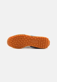 Suela de goma naranja con patrones de agarre circulares; diseño superior azul y blanco que proporciona contraste; presenta un logo de Puma.