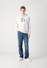 Sudadera blanca con un estampado gráfico en negro y amarillo, combinada con unos jeans de denim azul. El modelo lleva zapatillas blancas y está de pie sobre un fondo neutro.
