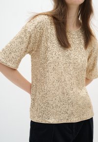 Femme portant un haut beige à manches courtes recouvert de sequins brillants et un pantalon noir, les mains derrière le dos.