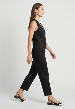 Vrouw die loopt in een zwarte mouwloze gebuttonde vest, bijpassende cropped broek en zwart-wit-gele puntige slingback schoenen.