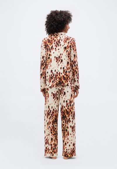 Γυναίκα με σκουρόχρωμα σγουρά μαλλιά, ντυμένη με κοστούμι κρεμ με καφέ και μαύρα ακανόνιστα στίγματα, που στέκεται γυρισμένη προς τα πίσω πάνω σε απλό φόντο.