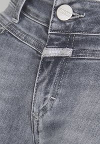 Detailní záběr na šedé džínové kalhoty ukazující kovový knoflík a látkovou visačku s nápisem "CLOSED" poblíž pásku a švu kapsy.