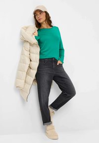 Femme appuyée contre un mur, portant une doudoune beige par-dessus un t-shirt vert à manches longues, un jean noir, des chaussettes beiges et des bottines beiges.