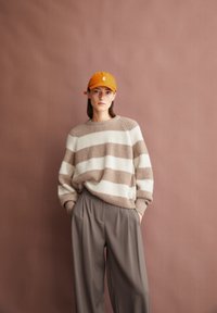 Pull en tricot rayé crème et marron clair, associé à un pantalon gris fluide. Le mannequin porte une casquette orange, sur fond rose.