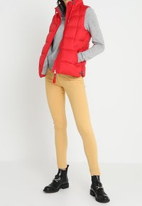 Rode puffy vest met hoge kraag, ritsluiting, gedragen over een grijze trui. Gecombineerd met lichtgele skinny broek en zwarte enkelboots.