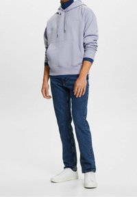 Sudadera gris clara con un bolsillo canguro, puños elásticos y dobladillo, combinada con jeans azules y zapatillas blancas. Diseño simple, estilo casual.