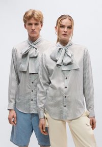 Ριγέ πουκάμισα με κουμπιά από ελαφρύ βαμβάκι, με υπερμεγέθεις παπιγιόν, καφέ κουμπιά και μανίκια που κάνουν ρολό, φορεμένα με τζιν σορτς και ανοιχτόχρωμο παντελόνι.