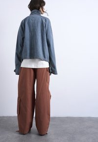 Γυναίκα που στέκεται σε τσιμεντένιο πάτωμα, φοράει μπλε τζιν μπουφάν, άσπρο πουκάμισο και φαρδιά καφέ παντελόνια cargo, κοιτώντας έναν λευκό τοίχο.