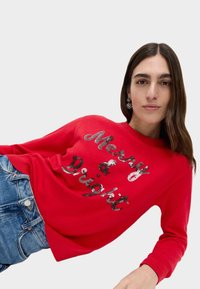 Maglione rosso lavorato a maglia con la scritta "Merry & Bright" in paillettes luccicanti. Maniche e orlo a costine, abbinato a jeans in denim blu. Tessuto liscio.