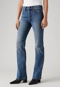 Personne portant un jean bleu taille haute à jambes droites, associé à des bottes noires à bout pointu et un haut blanc sur un fond uni.