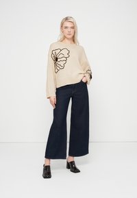 Béžový pletený svetr s hnědým květinovým vzorem, kombinovaný s tmavými džíny s širokými nohavicemi a černými kotníkovými botami na podpatku. Hladká textura.