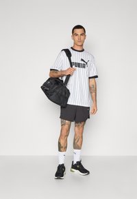Sort duffel bag, stribet hvid og sort t-shirt med logo, grå shorts, sorte sportssko og hvide sokker. Tatoveringer synlige på benene.