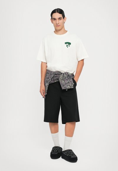 Camiseta branca com logotipo verde, calções pretos, faixa cinza com padrão à volta da cintura, meias brancas e sapatos pretos slip-on com design floral.