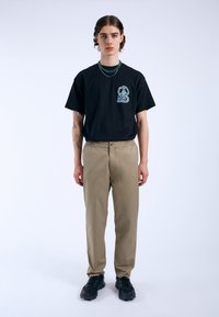 Zwarte T-shirt met korte mouwen en turquooise grafiek van een konijn op de linkerborst, gecombineerd met beige broek en zwarte sneakers, staand tegen een witte achtergrond.