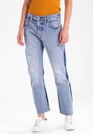 Person mit zweifarbigen, blauen Straight-Leg-Jeans mit Distressed-Details, ausgefransten Säumen, weißen Sneakern und einem senffarbenen, gerippten, ärmellosen Top.
