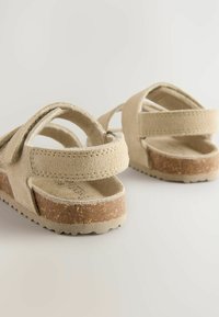 Beige Wildledersandalen mit Kreuzriemen, strukturierter Oberfläche und Korksohle. Offenes Rückendesign mit genähten Details und einem Rutschmuster.
