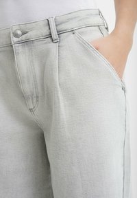 Jasnoszare dżinsy o luźnym kroju, z detalami takimi jak guzik i jedna kieszeń. Tkanina ma miękką strukturę z delikatnym przytłumieniem kolorów.