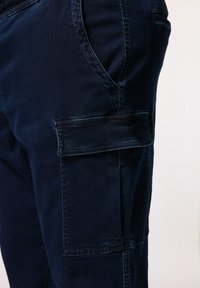 Granatowe spodnie cargo z bawełny, posiadające boczną kieszeń z klapką, gładką fakturę oraz standardowy pas ze szlufkami na pasek.