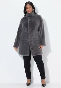 Abrigo de piel sintética gris con cuello alto, cierre de botones en la parte delantera y bolsillos laterales. Lleva puesto sobre pantalones y zapatos negros, con una textura suave.