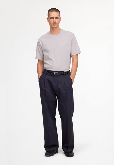 Homme debout, les mains dans les poches, portant un t-shirt gris clair, une ceinture noire, un pantalon ample foncé et des chaussures noires sur fond blanc.