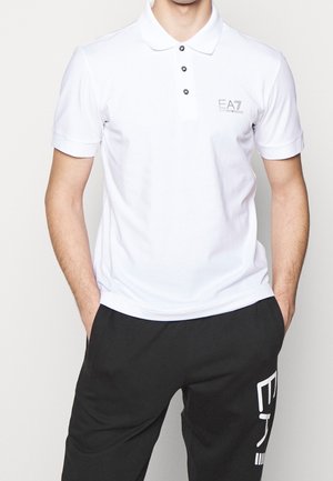 Mężczyzna noszący białą koszulkę polo EA7 Emporio Armani i czarne spodnie z białym napisem na lewej nodze, ręce w kieszeniach.