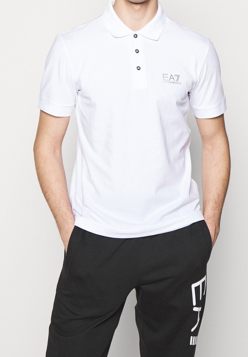 Homme portant un polo blanc EA7 Emporio Armani et un pantalon noir avec un motif texte blanc sur la jambe gauche, les mains dans les poches.